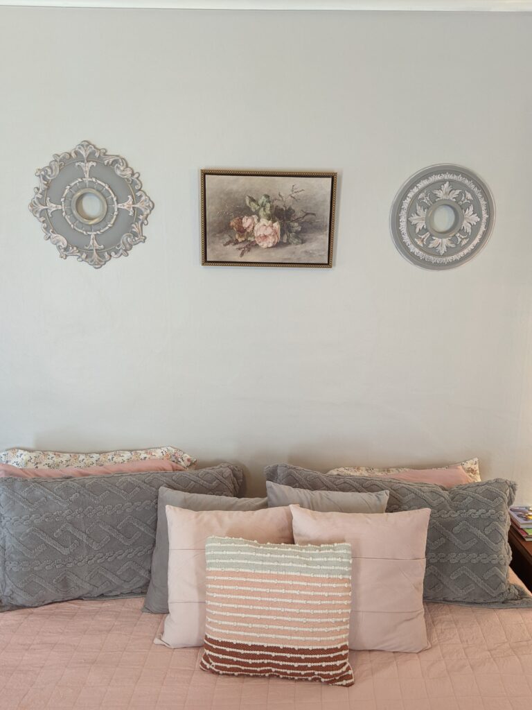 Instead of a headboard, the wall above the bed is decorated with painted vintage ceiling medallions and a piece of art. The color scheme is light pink and greay.
